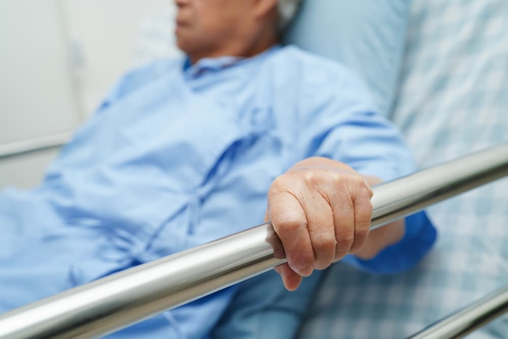 Elderly Asian woman patient lying in hospital bed, holding onto the bed rail, waiting for her family.