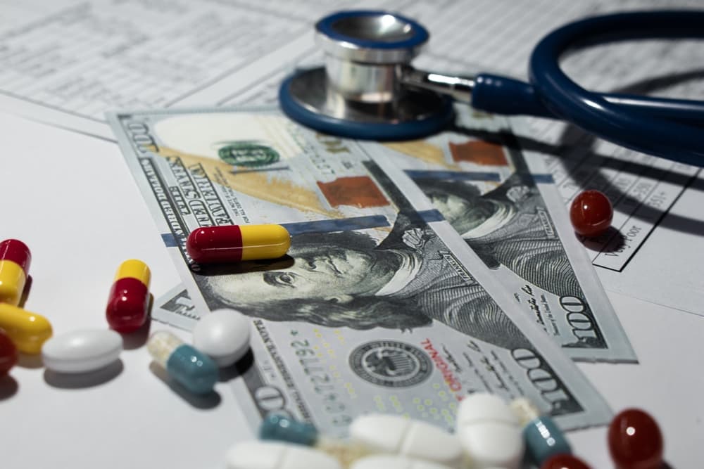 Assorted pills arranged on a one hundred dollar bill, symbolizing the cost of healthcare and medical treatment.
