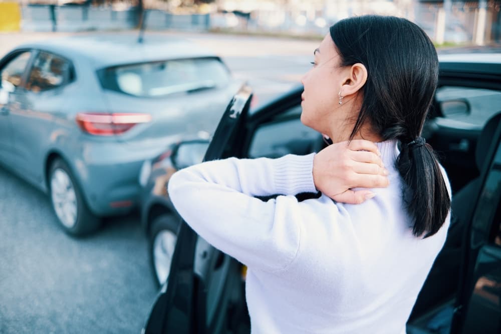 A woman holding her neck in pain after a rear-end collision, standing next to her car.