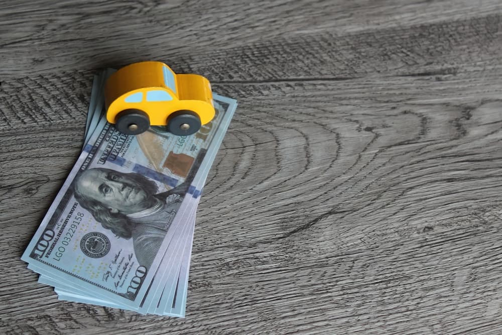 A stack of hundred-dollar bills topped with a small yellow toy car on a wooden surface.