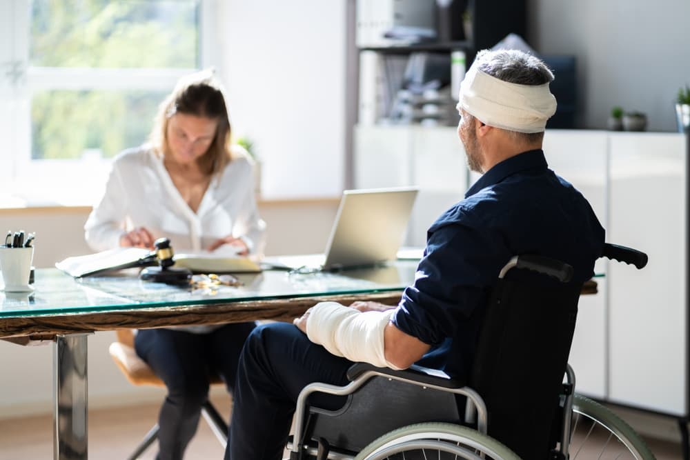 An injured man in a wheelchair with bandages on his head and arm consulting with a lawyer.