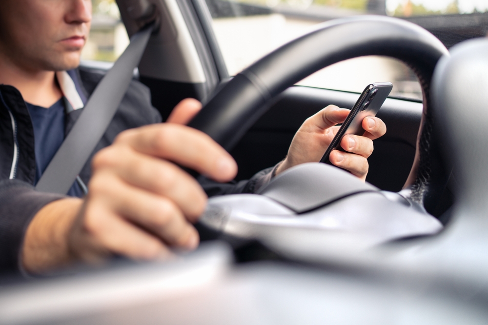 A driver holding a smartphone while steering a car, focusing on the phone screen.