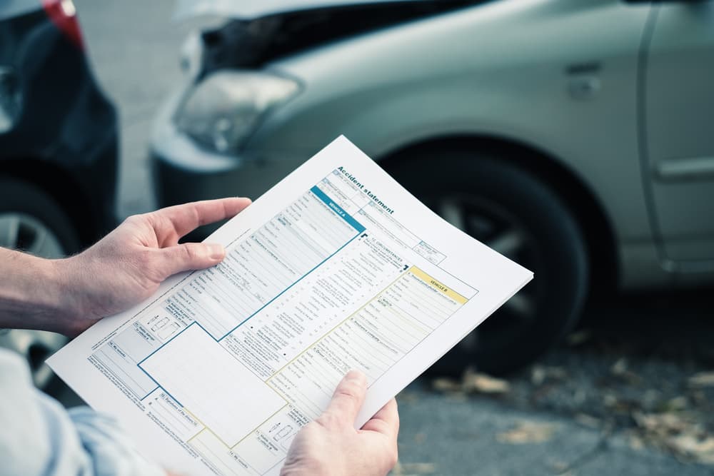 A person holding an accident statement form with damaged cars visible in the background.