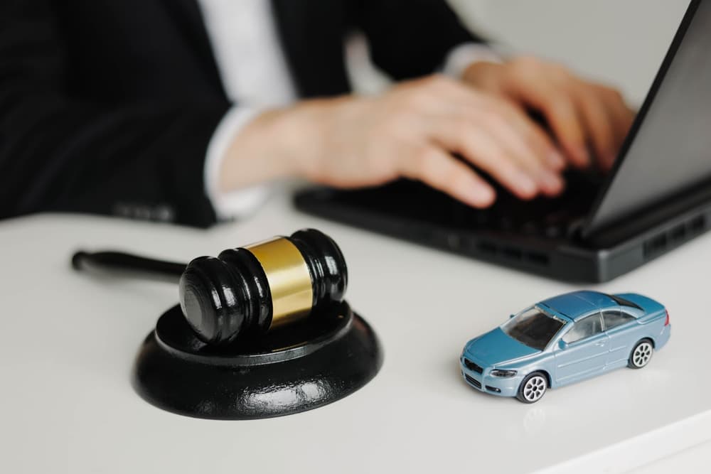 A gavel and a toy car on a desk with a person typing on a laptop.