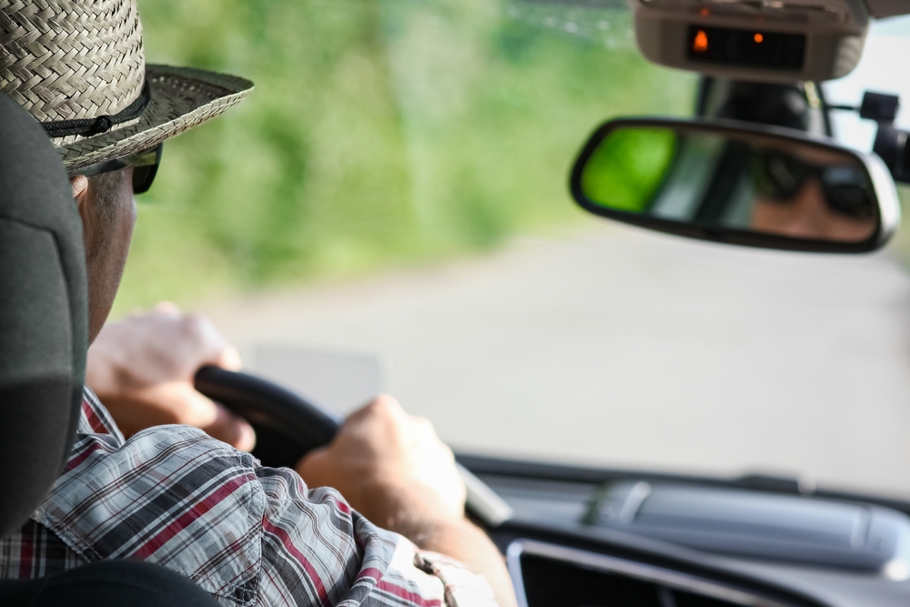 A man wearing a hat and sunglasses driving a car on a sunny day, seen from behind.