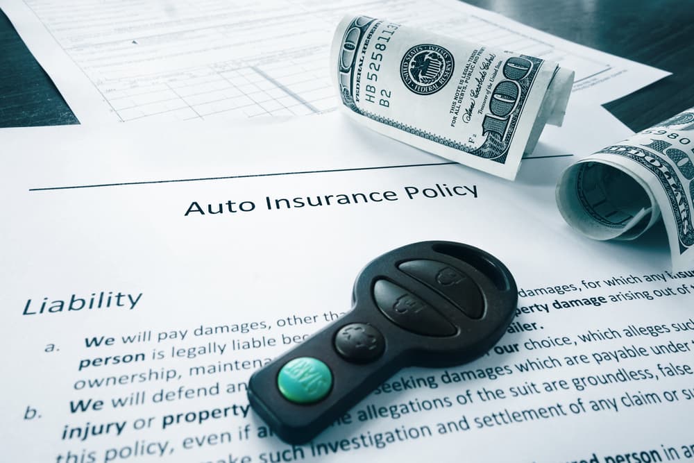 An auto insurance policy document with car keys and rolled-up hundred-dollar bills on top.