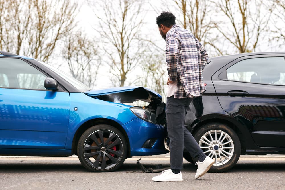 A man inspecting the damage to a blue car after a rear-end collision with a black car.