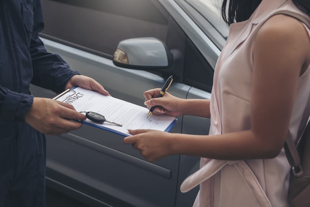 A woman signing a contract on a clipboard held by a man beside a car.