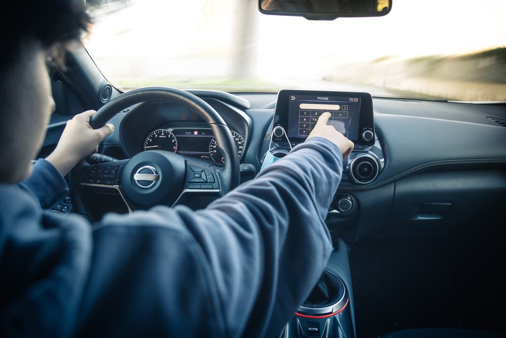 Teen driver uses infotainment, reading messages and calling while driving, risking accidents. Focus on hand, highlighting youth and distraction.
