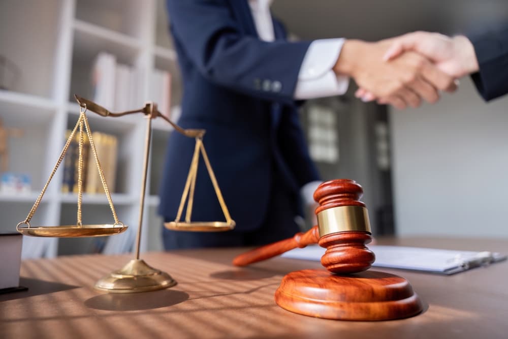Lawyer handshake at client meeting with scales of justice and gavel on desk, symbolizing legal agreement in a modern office.